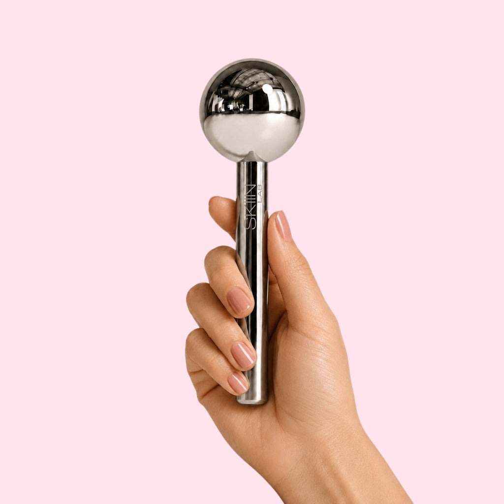 Main tenant le Cryo Ice Glow SkiinLAB par son manche cylindrique en acier chromé gravé SKIIN LAB, sphère polie miroir orientée vers le haut, ongles en manucure claire, fond rose uni